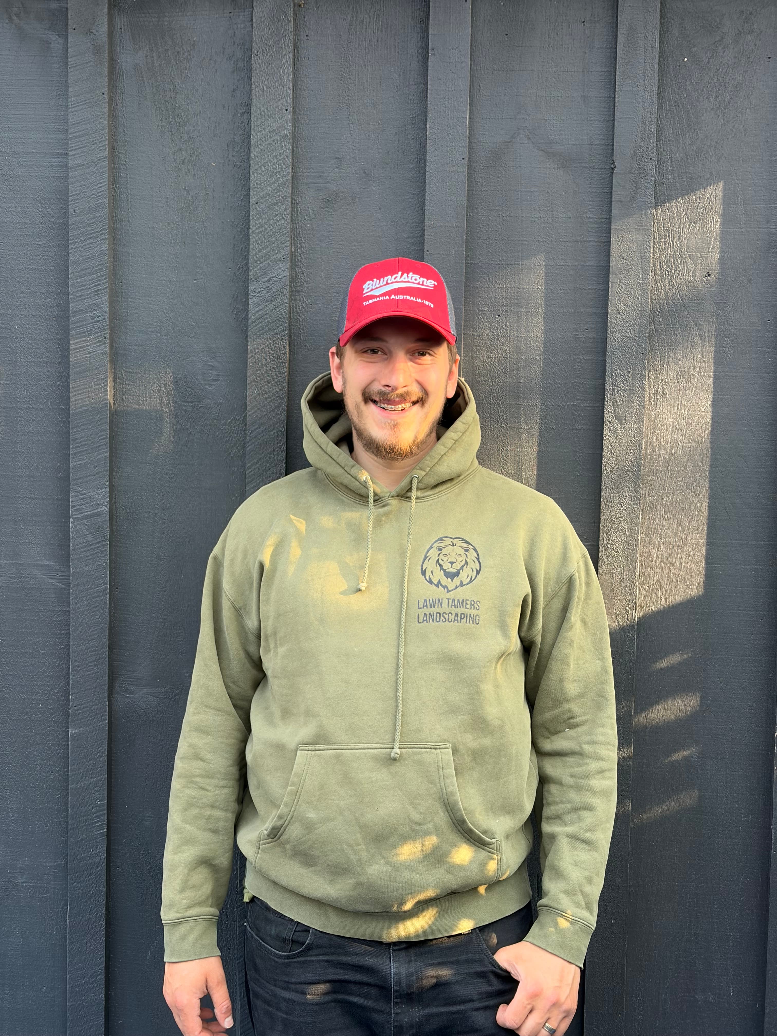Smiling man wearing a red cap and green hoodie against a dark wooden wall.