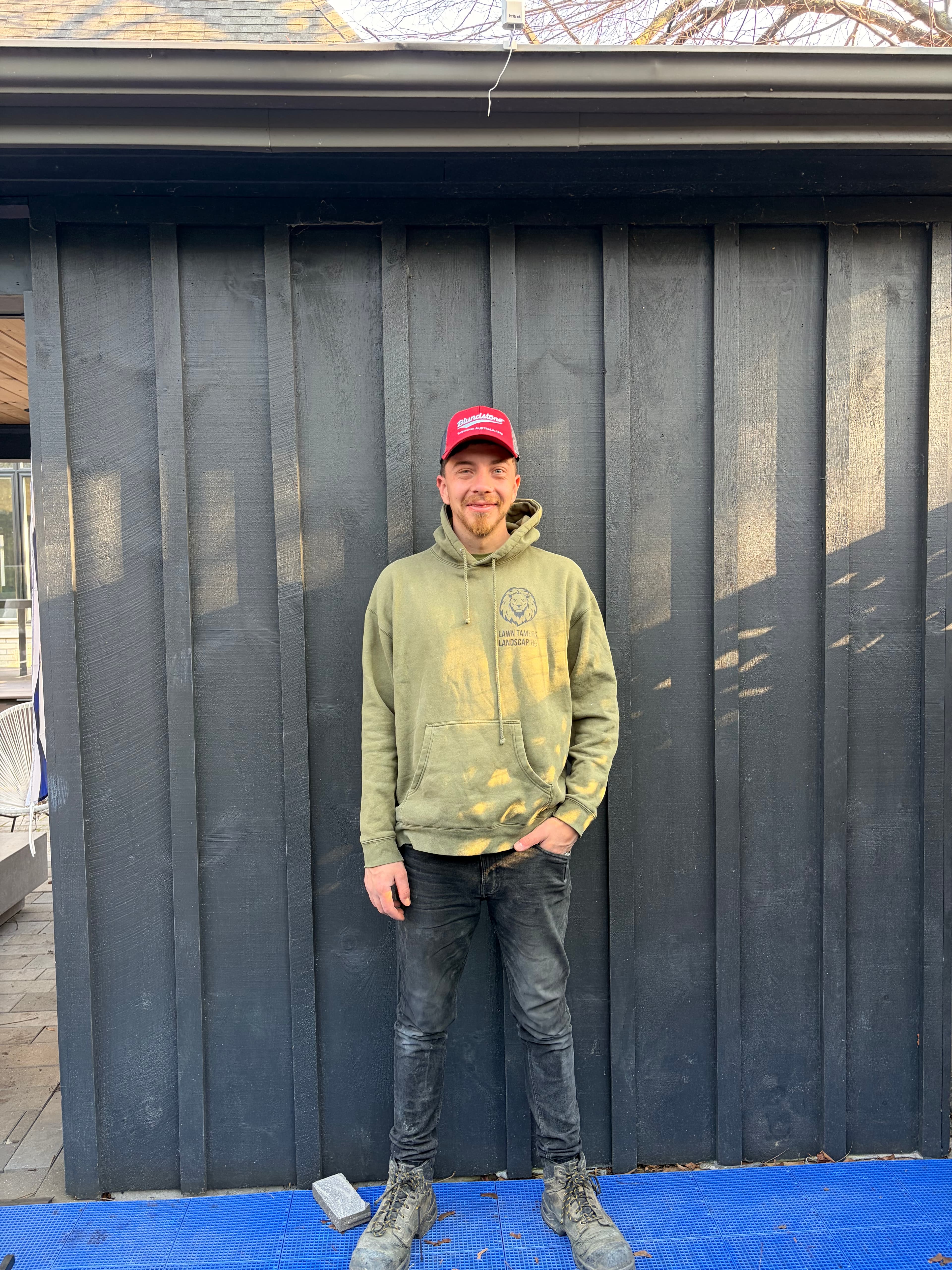Smiling man in a red cap and green hoodie against a dark wooden wall.